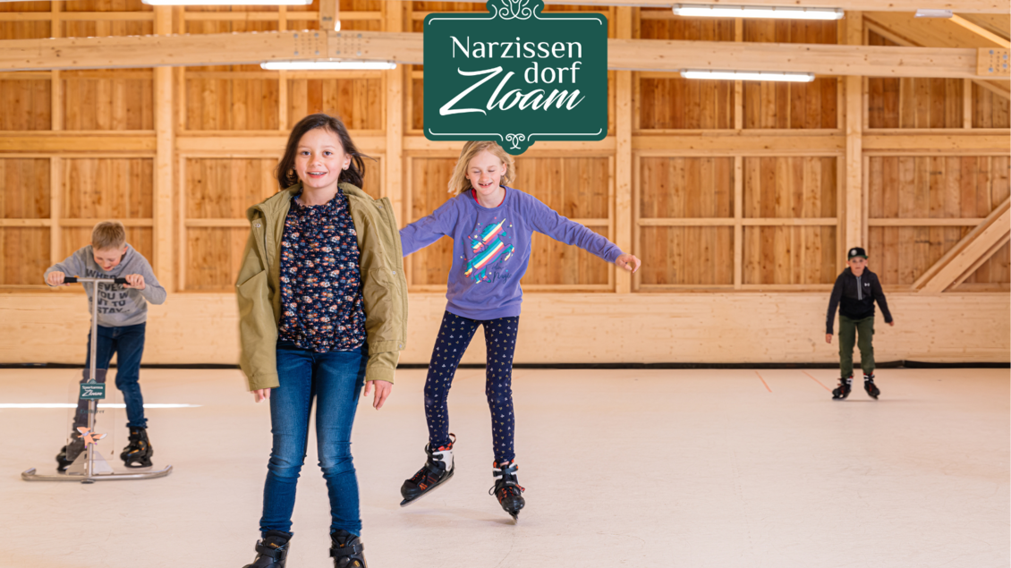 Ein Raum mit Kindern, die auf Schlittschuhen fahren. Im Hintergrund ist ein Holzinterieur zu sehen, und das Logo "Narzissen Dorf Zloam" ist oben angebracht. | © Narzissendorf Zloam, www.zloam.at