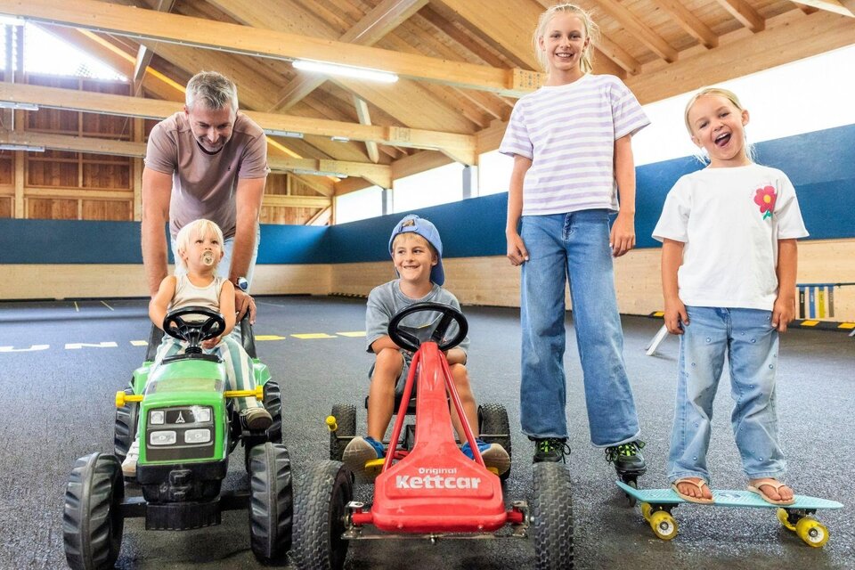 Eine fröhliche Gruppe von Kindern fährt mit Spielzeugfahrzeugen in einer hellen Indoor-Spielumgebung. Ein Erwachsener steht hinter den fahrbaren Geräten und lächelt. | © www.zloam.at