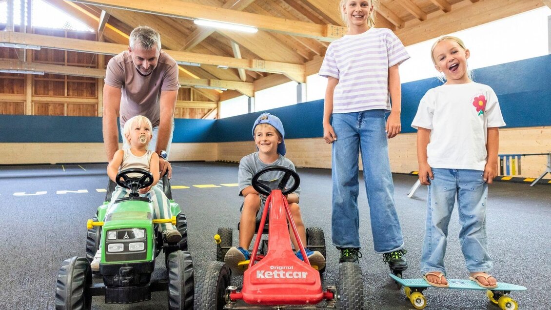 Eine fröhliche Gruppe von Kindern fährt mit Spielzeugfahrzeugen in einer hellen Indoor-Spielumgebung. Ein Erwachsener steht hinter den fahrbaren Geräten und lächelt. | © www.zloam.at