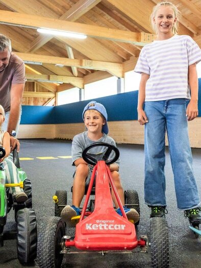 A cheerful group of children is riding toy vehicles in a bright indoor play environment. An adult stands behind the moving devices and smiles. | © www.zloam.at