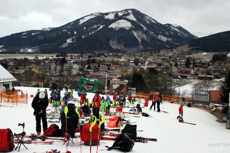 Eine belebte Skistation mit zahlreichen Skifahrern und vielen Skiausrüstungen auf dem Schnee. Im Hintergrund sind Berge und eine malerische Stadt zu sehen. | © WSV Raiffeisen Turnau
