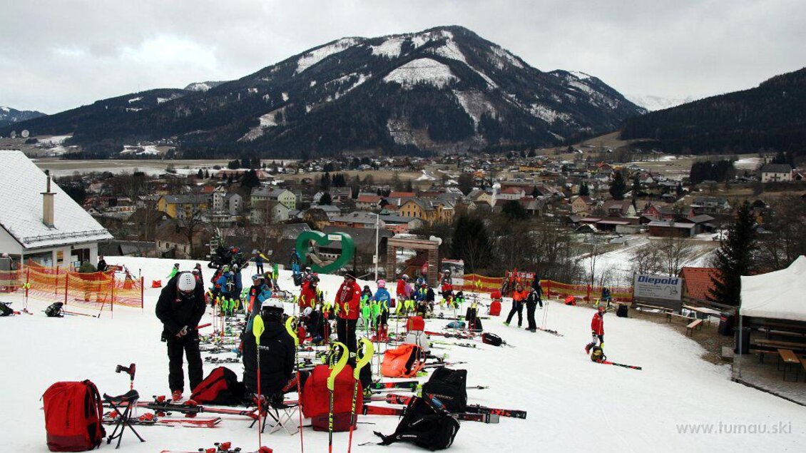 Eine belebte Skistation mit zahlreichen Skifahrern und vielen Skiausrüstungen auf dem Schnee. Im Hintergrund sind Berge und eine malerische Stadt zu sehen. | © WSV Raiffeisen Turnau