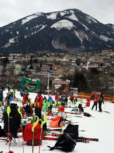 Eine belebte Skistation mit zahlreichen Skifahrern und vielen Skiausrüstungen auf dem Schnee. Im Hintergrund sind Berge und eine malerische Stadt zu sehen. | © WSV Raiffeisen Turnau