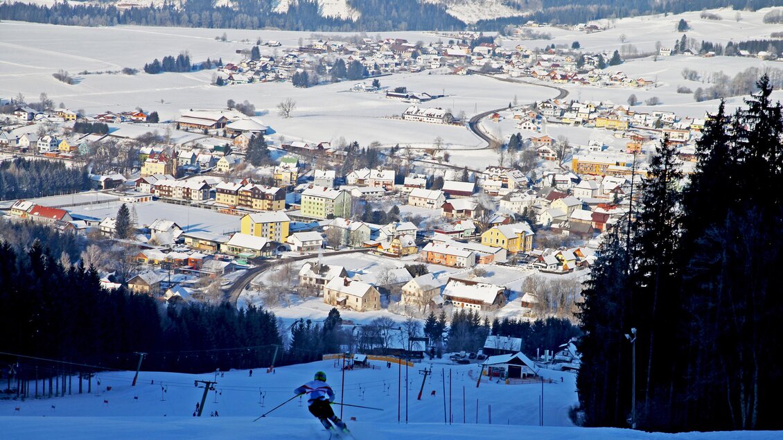 Eine schneebedeckte Landschaft mit einem Skifahrer im Vordergrund. Im Hintergrund ist ein malerisches Dorf zu sehen, umgeben von verschneiten Bergen. | © Günther Breidler