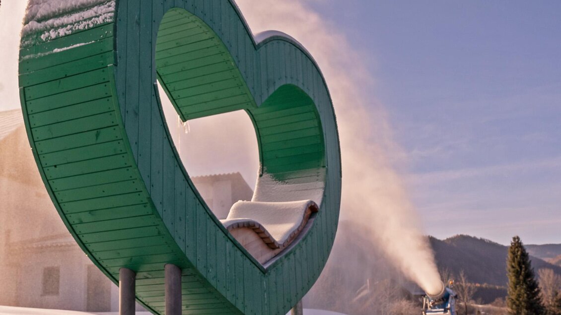Ein grünes Herz aus Holz im Schnee. Im Hintergrund wird Kunstschnee erzeugt. | © Günther Breidler
