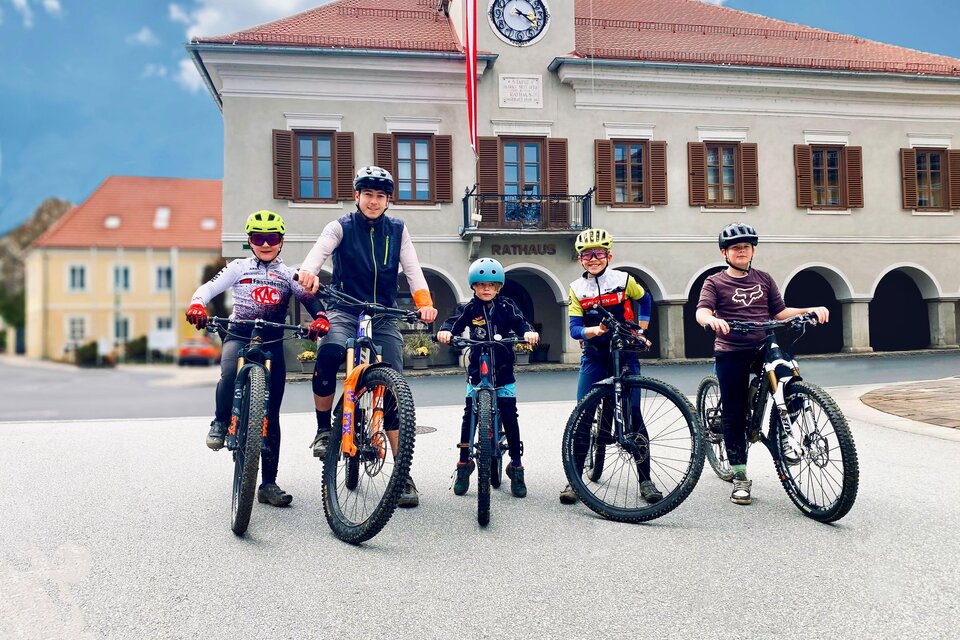 Eine Gruppe von fünf Kindern steht mit ihren Fahrrädern auf einer Straße vor einem Gebäude. Sie tragen Helme und Fahrradausrüstung, und der Himmel ist blau. | © TV Südsteiermark - AndreasMühlbacher im.puls sport