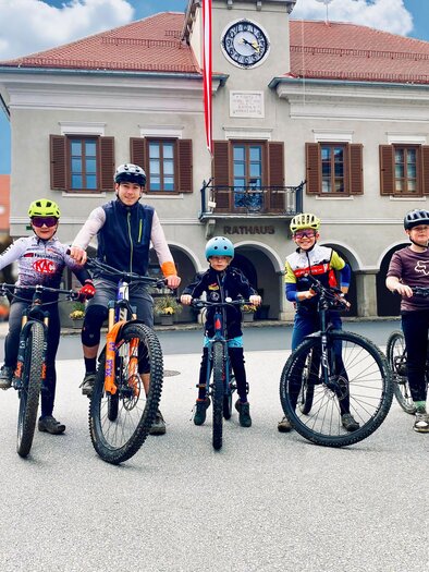 A group of five children stands with their bicycles on a street in front of a building. They are wearing helmets and cycling gear, and the sky is blue. | © TV Südsteiermark - AndreasMühlbacher im.puls sport