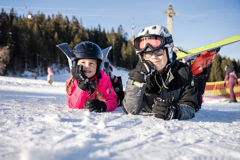 Zwei Kinder liegen im Schnee und lächeln. Sie tragen Skikleidung und Helme, während im Hintergrund Bäume und eine Skilift sichtbar sind. | © Tourismusverband Oststeiermark