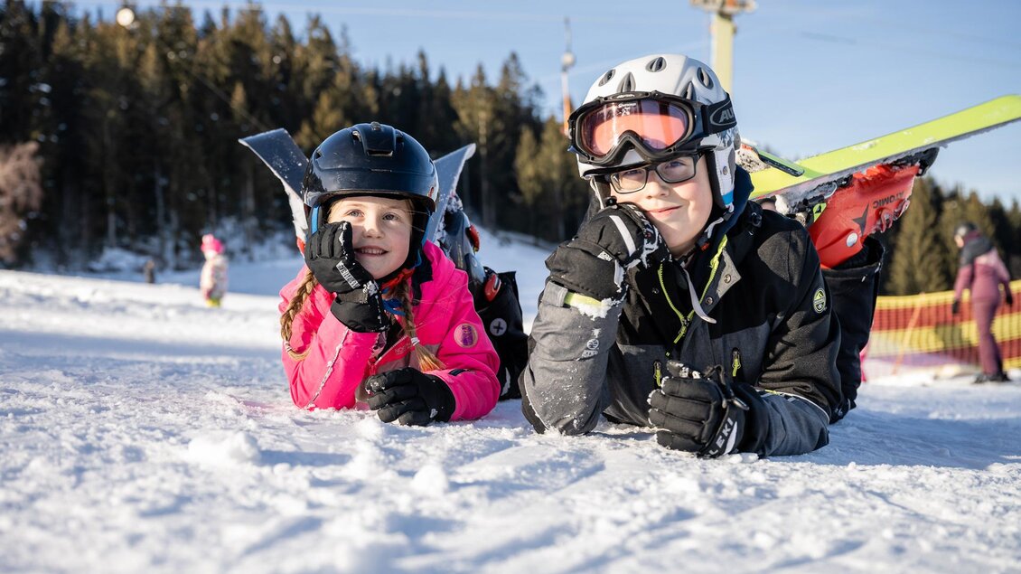 Zwei Kinder liegen im Schnee und lächeln. Sie tragen Skikleidung und Helme, während im Hintergrund Bäume und eine Skilift sichtbar sind. | © Tourismusverband Oststeiermark