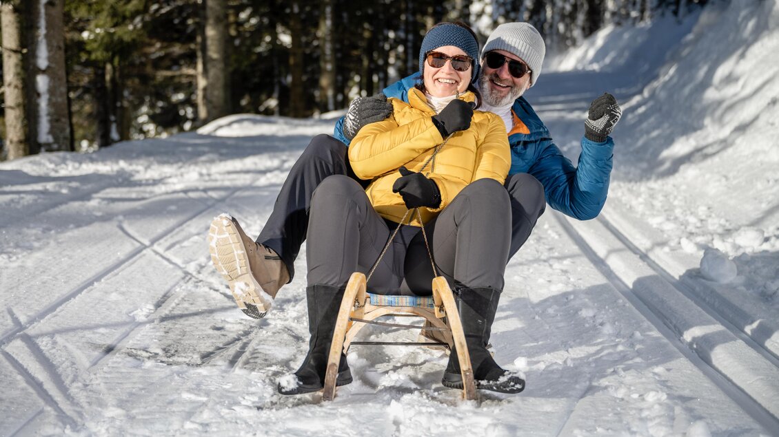 Zwei Personen sitzen fröhlich auf einem Schlitten im Schnee. Die Bäume im Hintergrund sind mit Schnee bedeckt und es scheint sonnig zu sein. | © Tourismusverband Oststeiermark