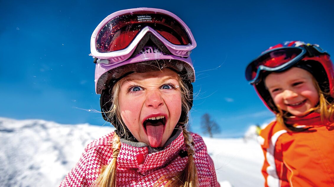 Ein fröhliches Kind mit Skibrille macht eine lustige Grimasse im Schnee. Im Hintergrund ist ein anderes Kind zu sehen, das ebenfalls Spaß hat. | © Almblick Skilift