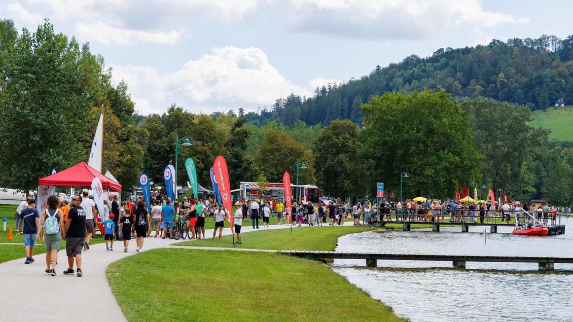 Eine belebte Promenade am See mit vielen Besuchern und bunten Fahnen. Im Hintergrund sind Bäume und ein Hügel sichtbar. | © Woche Familienfest