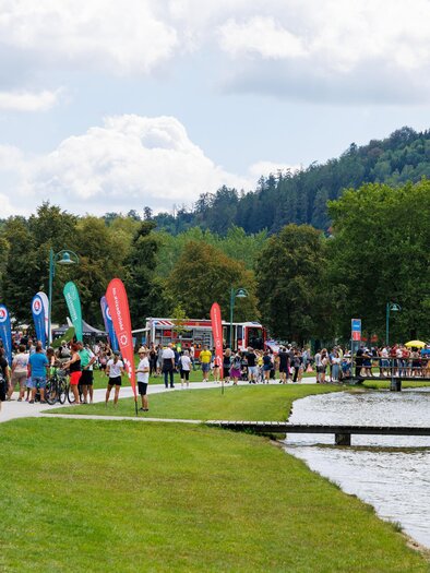A bustling promenade by the lake with many visitors and colorful flags. In the background, trees and a hill are visible. | © Woche Familienfest