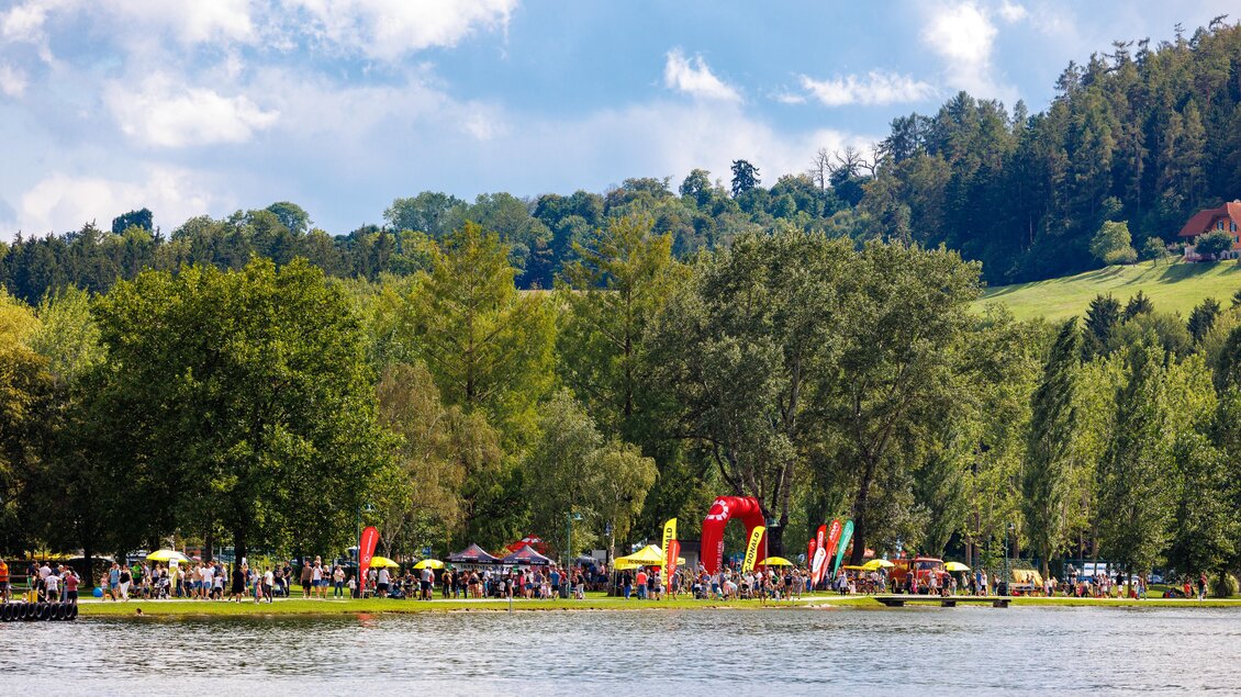 Ein malerischer See umgeben von Bäumen und blühenden Wiesen. Am Ufer sind Menschen mit bunten Flaggen und Sonnenschirmen versammelt. | © Woche Familienfest