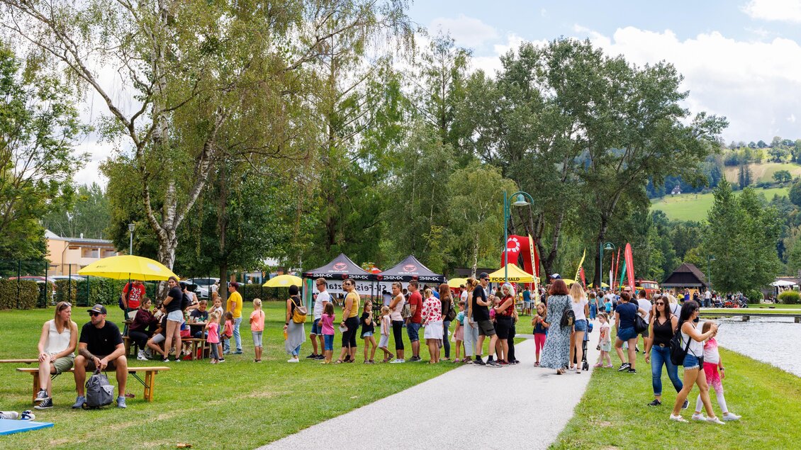 Eine belebte Parklandschaft mit vielen Menschen, die entlang eines Weges und eines Gewässers stehen. Bäume und gelbe Sonnenschirme sorgen für eine angenehme Atmosphäre. | © Woche Familienfest