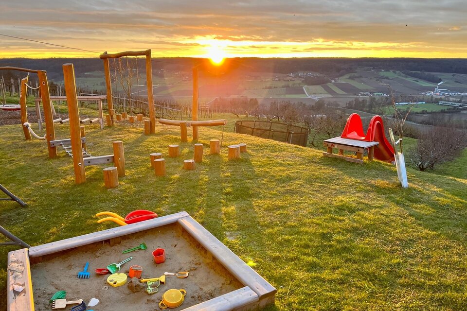 Ein Spielplatz mit einer Sandkiste, einer Rutsche und Schaukeln liegt auf einer Wiese. Im Hintergrund strahlt die Sonne bei Sonnenuntergang über die sanften Hügel. | © Maluhof Lugitsch Markus
