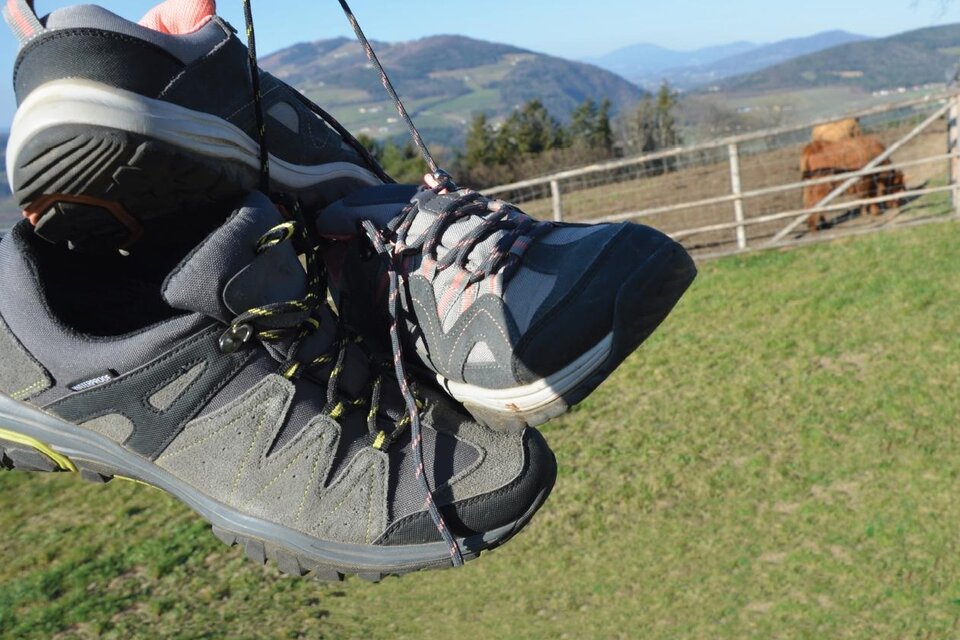 Ein paar Wanderschuhe hängen an einem Schnürsenkel. Im Hintergrund sind grüne Wiesen und sanfte Hügel zu sehen. | © Doris Allmer