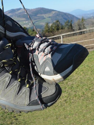 Ein paar Wanderschuhe hängen an einem Schnürsenkel. Im Hintergrund sind grüne Wiesen und sanfte Hügel zu sehen. | © Doris Allmer