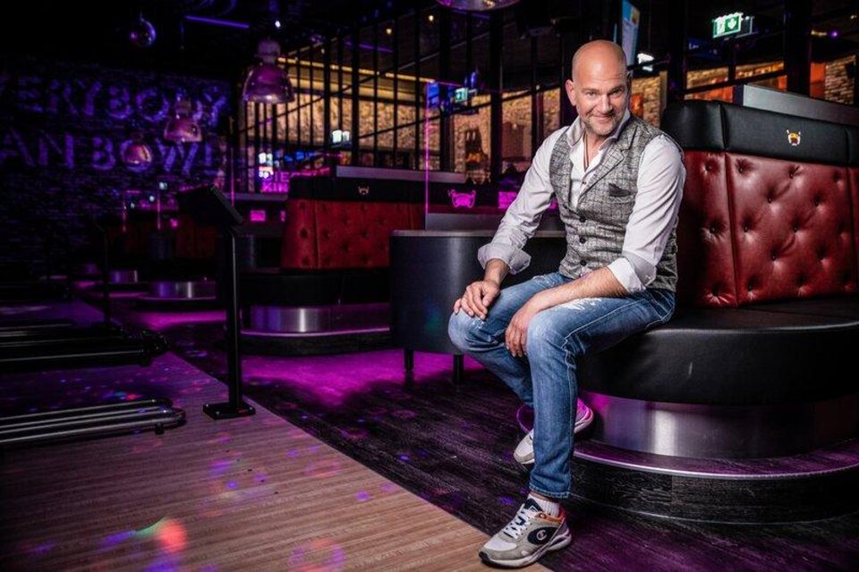 Ein Mann sitzt lächelnd auf einer gepolsterten Bank in einem modernen Raum. Die Umgebung ist dunkel mit bunten Lichtern und einer lebhaften Atmosphäre. | © Udo Wenders