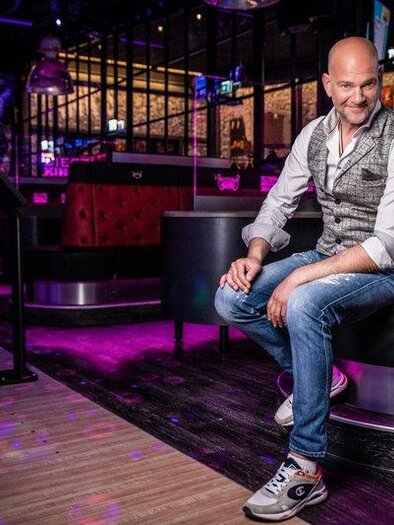 Ein Mann sitzt lächelnd auf einer gepolsterten Bank in einem modernen Raum. Die Umgebung ist dunkel mit bunten Lichtern und einer lebhaften Atmosphäre. | © Udo Wenders