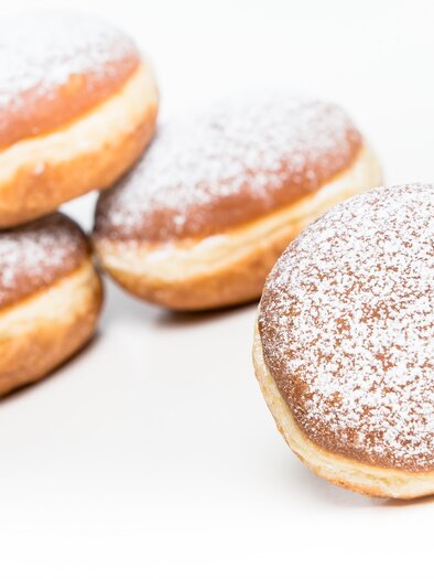Egy csoport ízletes, porcukorral megszórt fánk. A fánkok aranybarna színűek és egy fehér háttéren helyezkednek el. | © Bäckerei Felber
