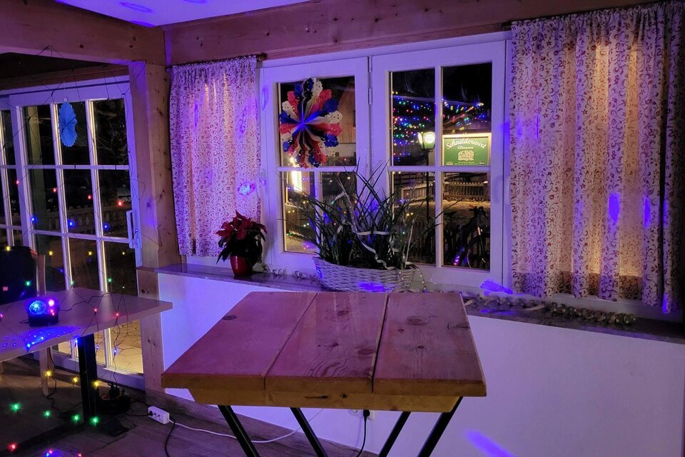 Ein gemütlicher Raum mit einem Holztisch und bunten Lichtern. Die Fenster sind mit Vorhängen geschmückt und Pflanzen stehen auf der Fensterbank. | © Petra Kirchschlager