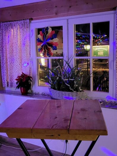 Ein gemütlicher Raum mit einem Holztisch und bunten Lichtern. Die Fenster sind mit Vorhängen geschmückt und Pflanzen stehen auf der Fensterbank. | © Petra Kirchschlager