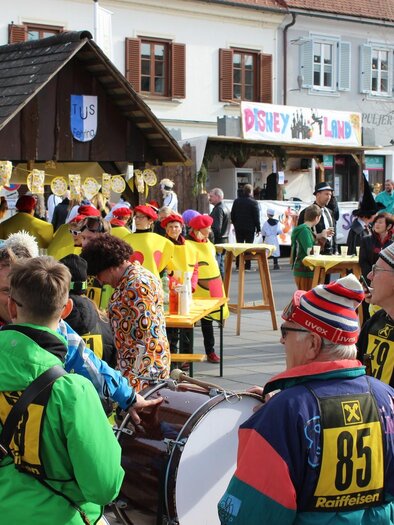 Eine bunte Menschenmenge versammelt sich auf einer Straße, während sie sich auf ein Fest vorbereitet. Lebhafte Kostüme und musikalische Elemente schaffen eine fröhliche Atmosphäre. | © Stadtgemeinde Fehring