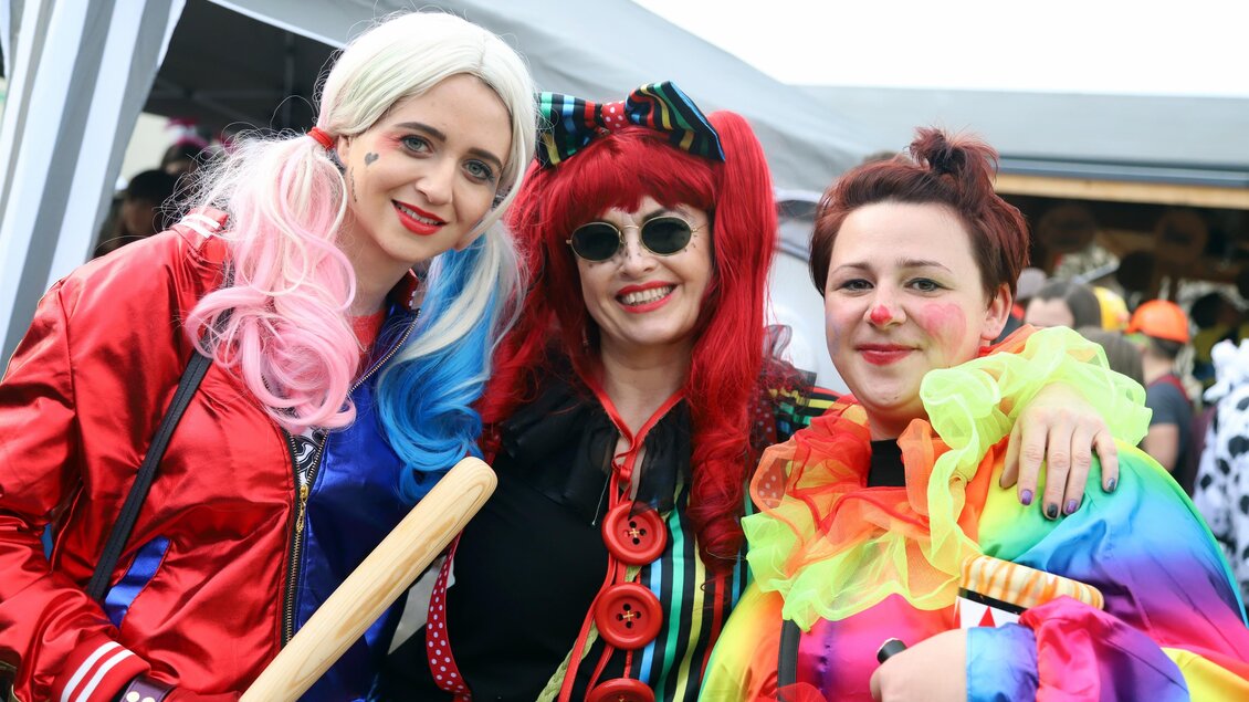 Drei Frauen in bunten Karnevalskostümen lächeln fröhlich in die Kamera. Sie tragen auffällige Perücken und Accessoires und scheinen Spaß zu haben.