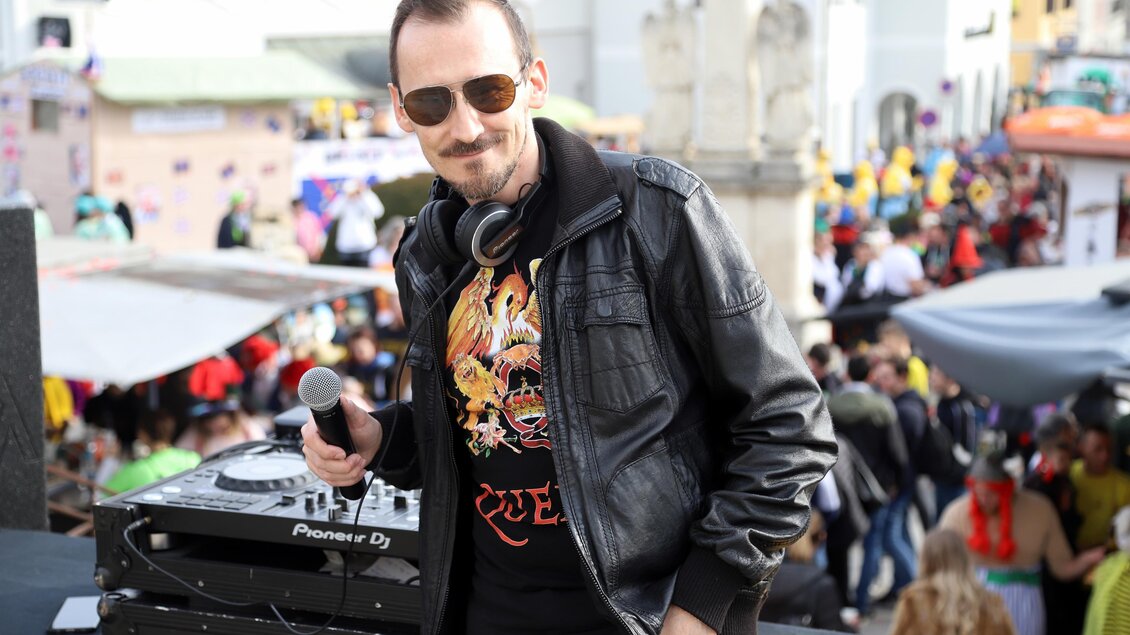 Ein DJ steht an einem Mischpult und hält ein Mikrofon in der Hand. Im Hintergrund sind viele Menschen auf einer Feier zu sehen. | © Markus Weiss