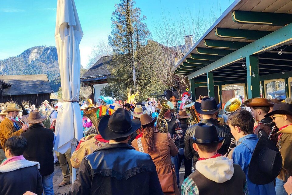 Eine Gruppe von Menschen in bunten Hüten und Kostümen versammelt sich im Freien. Im Hintergrund sind Bäume und Gebäude zu sehen, vor einer bergigen Kulisse. | © Petra Kirchschlager