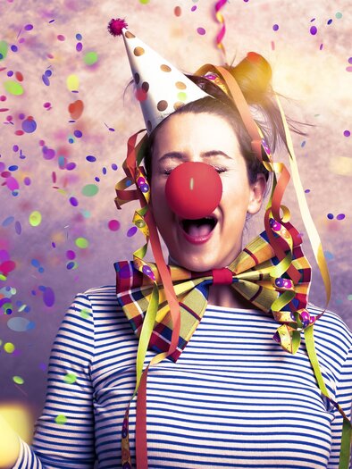 A cheerful person with a clown nose and a party hat stands in the midst of confetti. They are smiling and have colorful balloons around them. | © TV Südsteiermark - Drubig-Photo