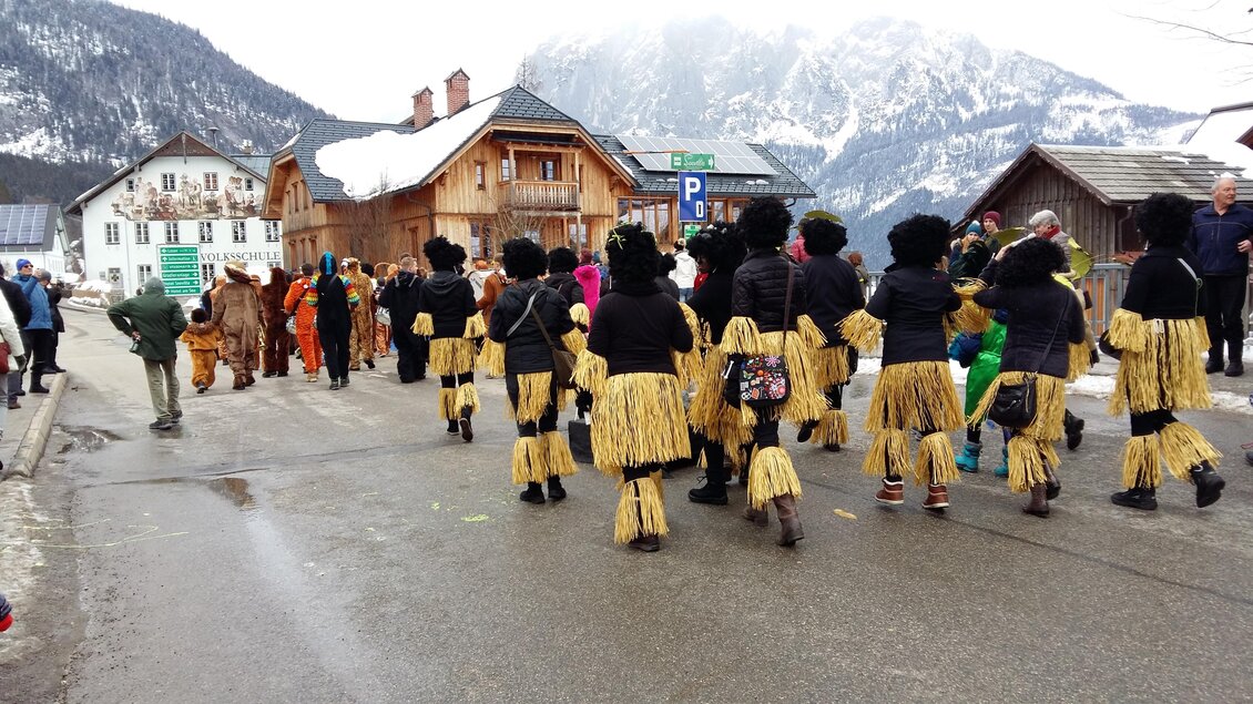 Eine Gruppe von Menschen in bunten Kostümen marschiert auf einer Straße. Im Hintergrund sind Berge und traditionelle Gebäude sichtbar. | © Kirchschlager Petra