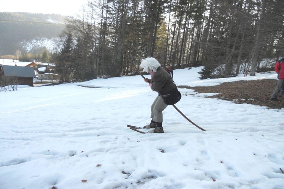Eine Person mit einer weißen Perücke fährt auf einem Ski durch den Schnee. Im Hintergrund sind Bäume und einige Hütten zu sehen. | © Kirchschlager Petra