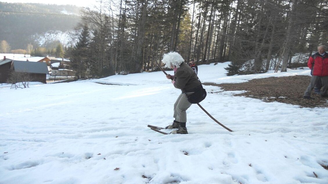 Eine Person mit einer weißen Perücke fährt auf einem Ski durch den Schnee. Im Hintergrund sind Bäume und einige Hütten zu sehen. | © Kirchschlager Petra