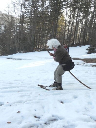 Eine Person mit einer weißen Perücke fährt auf einem Ski durch den Schnee. Im Hintergrund sind Bäume und einige Hütten zu sehen. | © Kirchschlager Petra