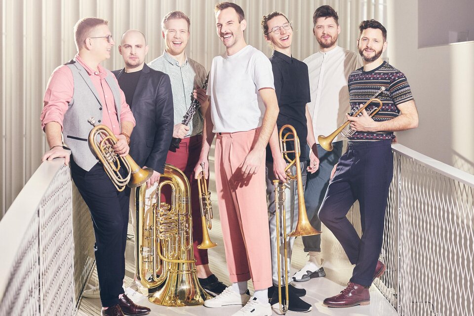 Eine Gruppe von sieben Musikern steht auf einer Bühne. Sie halten verschiedene Blasinstrumente und lachen freundlich in die Kamera. | © Felix Groteloh