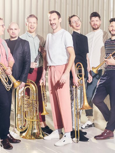 A group of seven musicians stands on a stage. They hold various wind instruments and smile kindly at the camera. | © Felix Groteloh