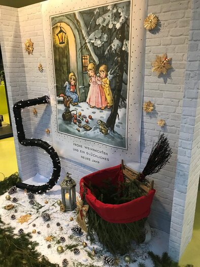 Ein weihnachtlich dekorierter Raum mit einer großen Wandmalerei von zwei Mädchen im Schnee. Im Vordergrund steht ein roter Korb, umgeben von Tannenzweigen und einer großen Zahl fünf. | © Stadtgemeinde Fehring