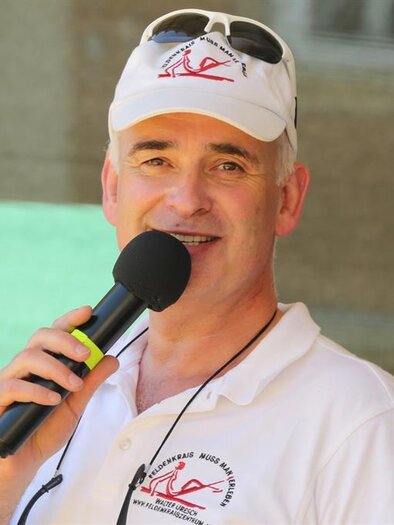 Ein Mann spricht mit einem Mikrofon und lächelt. Er trägt ein weißes T-Shirt und eine Mütze. | © Walter Uresch