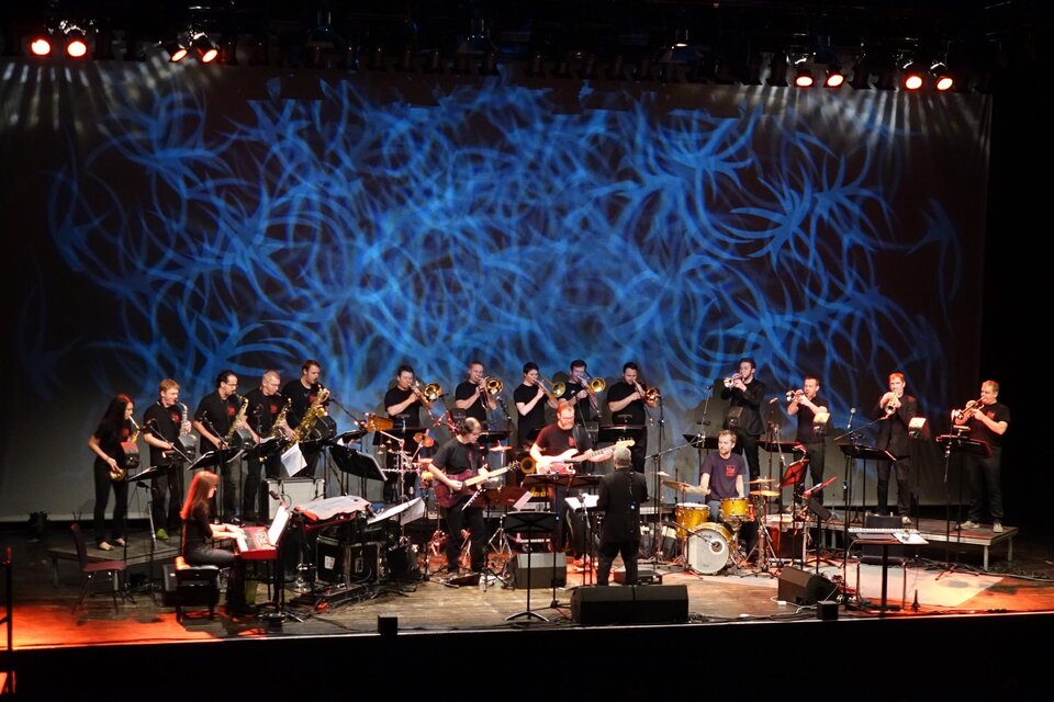 Eine große Band spielt auf der Bühne vor einem bunten Hintergrund. Die Musiker arbeiten gemeinsam an ihren Instrumenten und schaffen eine lebendige Atmosphäre.