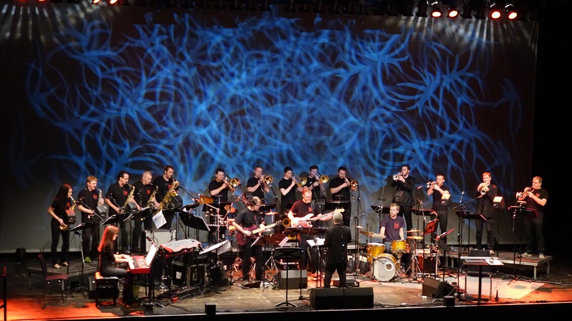 Eine große Band spielt auf der Bühne vor einem bunten Hintergrund. Die Musiker arbeiten gemeinsam an ihren Instrumenten und schaffen eine lebendige Atmosphäre.