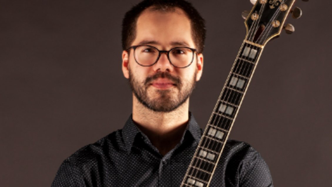 Ein Mann mit einer Gitarre posiert vor einem neutralen Hintergrund. Er trägt ein schwarzes Hemd mit Punkten und hat eine Brille auf. | © ©Emiliano Sampaio