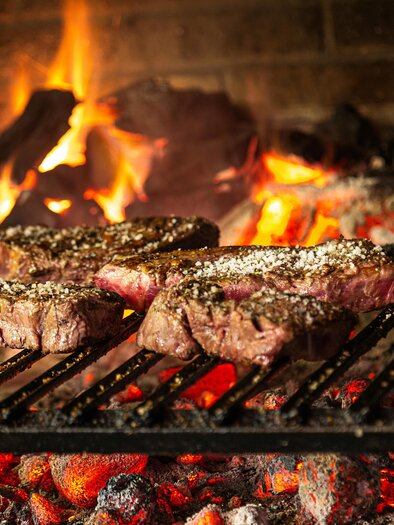 Juicy steaks sizzling over an open flame. In the background, the flames are flickering and the coals are glowing. | © Naturwelten Steiermark