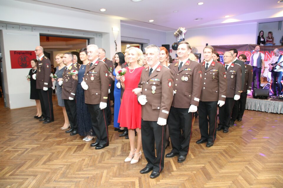 Eine Gruppe von uniformierten Personen steht lächelnd zusammen. Im Vordergrund ist eine Frau in einem roten Kleid zu sehen.