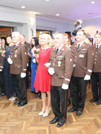 A group of uniformed individuals is smiling together. In the foreground, there is a woman in a red dress.