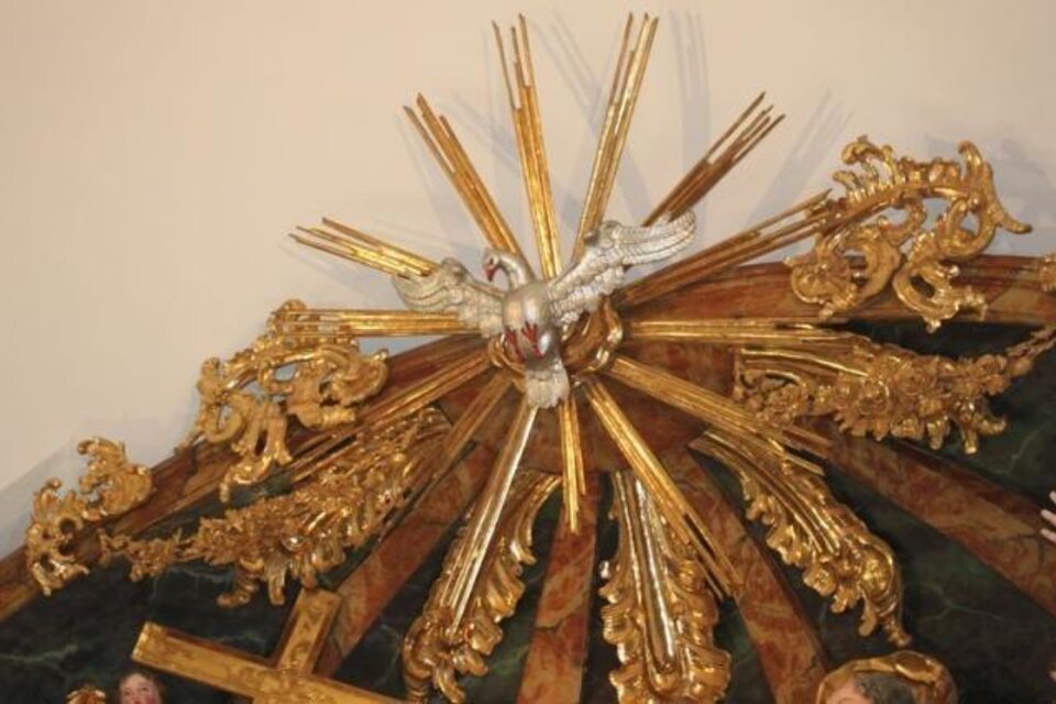 Ein kunstvolles goldenes Schmuckstück mit einem Vogel in der Mitte und strahlenden Lichtstrahlen. Es befindet sich an der Decke und verleiht dem Raum eine feierliche Atmosphäre. | © Pfarrkirche Anger