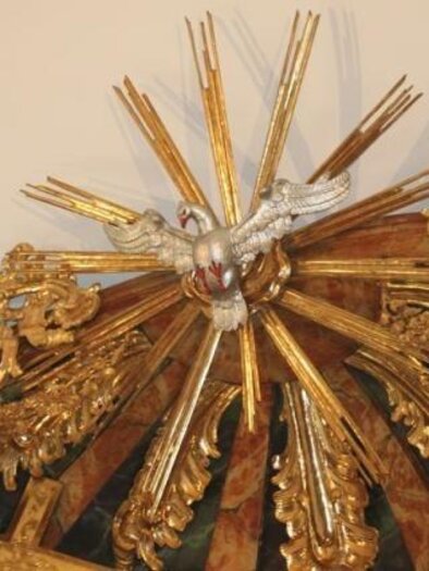 Ein kunstvolles goldenes Schmuckstück mit einem Vogel in der Mitte und strahlenden Lichtstrahlen. Es befindet sich an der Decke und verleiht dem Raum eine feierliche Atmosphäre. | © Pfarrkirche Anger