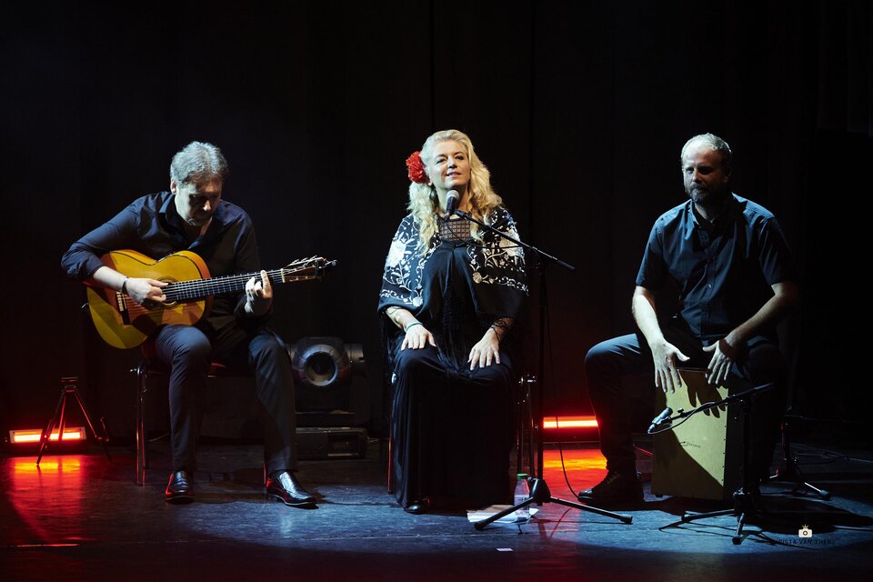 Trio auf dunkler Bühne: links ein Gitarrist im Sitzen, in der Mitte eine Sängerin am Mikrofon mit roter Blume im Haar, rechts ein Percussionist auf einer Cajón; dezentes rotes und blaues Bühnenlicht am Boden. | © Steckl