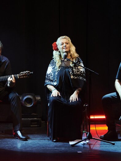 Trio auf dunkler Bühne: links ein Gitarrist im Sitzen, in der Mitte eine Sängerin am Mikrofon mit roter Blume im Haar, rechts ein Percussionist auf einer Cajón; dezentes rotes und blaues Bühnenlicht am Boden. | © Steckl